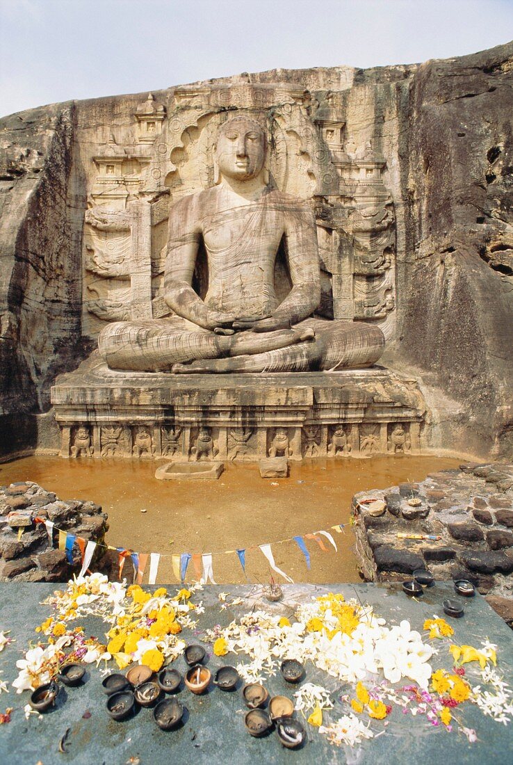 In Fels gehauene, sitzende Buddhastatue mit davor liegenden Opfergaben; Polonnaruwa, Sri Lanka