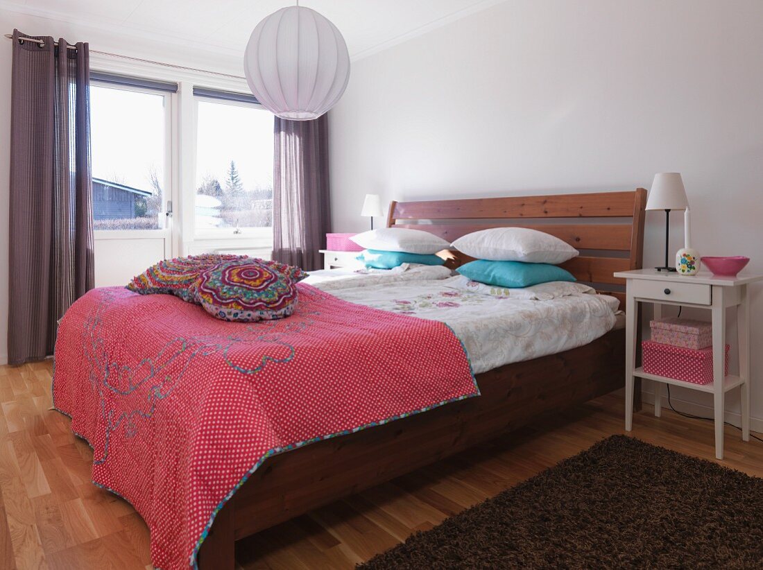 Bed with solid wooden frame and headboard and colourful bedspread and pillows next to balcony door in bedroom
