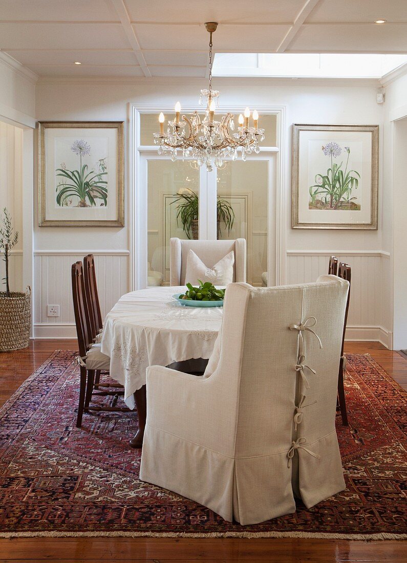 Armchair with pale loose cover, chairs around table with white tablecloth and chandelier in traditional, elegant dining room