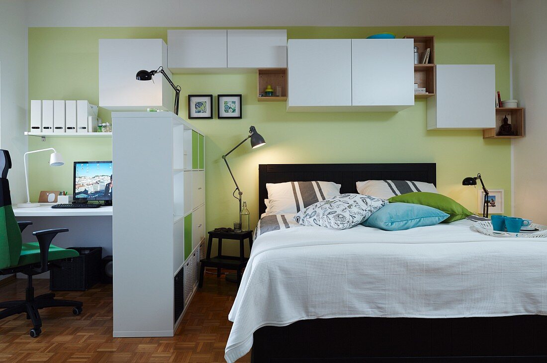 A room divider and wall cupboards hung at different heights to create lots of storage space between a double bed and a home office with a communal, green-painted wall