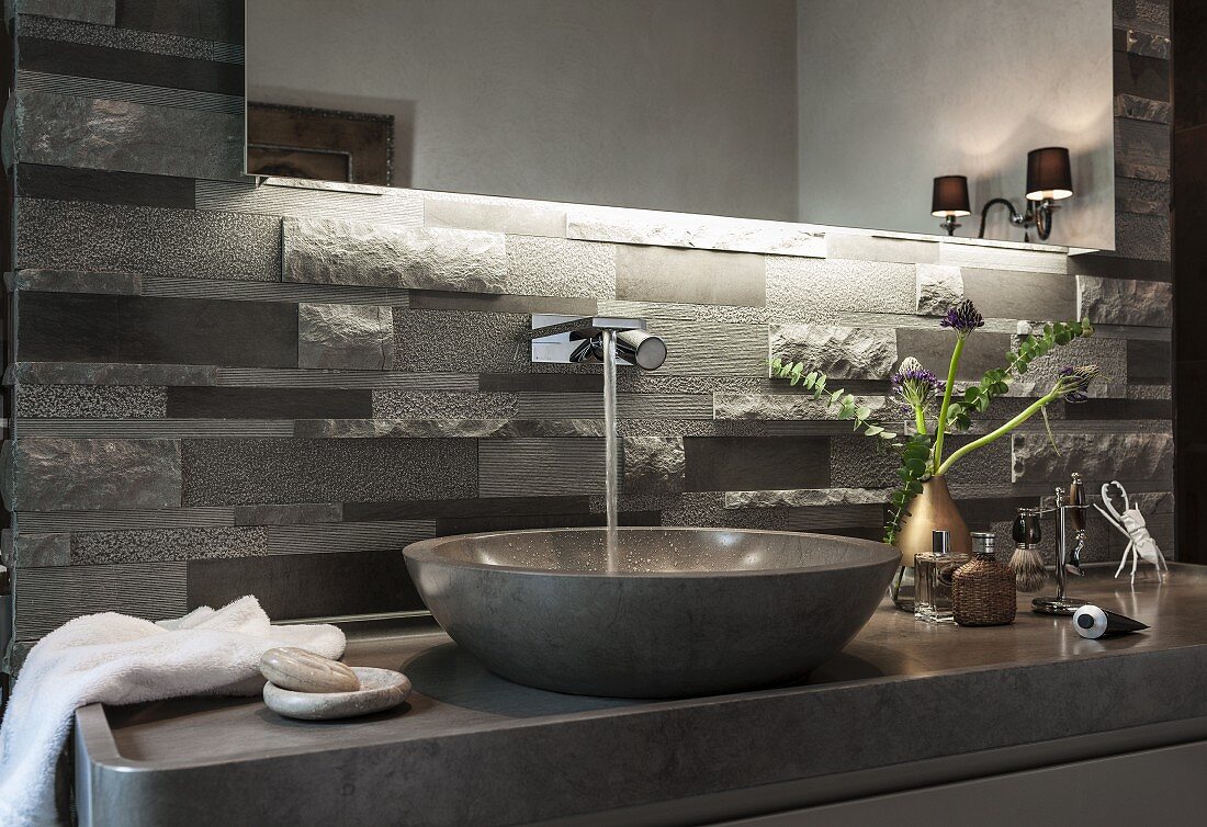 An illuminated mirror above polished limestone washstand with a console basin and grey limestone relief tiles