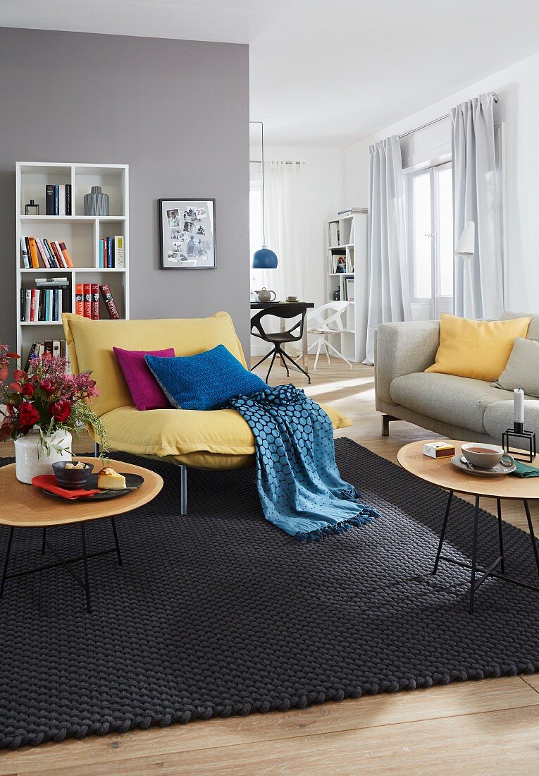 A grey living room with a yellow armchair with a throw and cushions and a white bookshelf