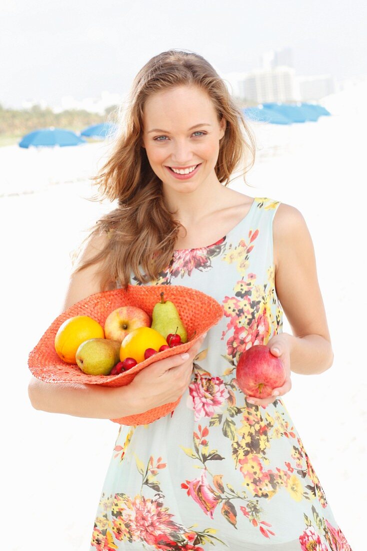 A young woman wearing a summer dress holding a hat filled with fruit and holding out an apple