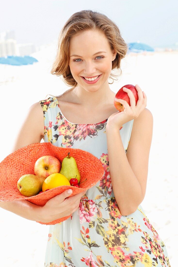 A young woman wearing a summer dress holding a hat filled with fruit and an apple