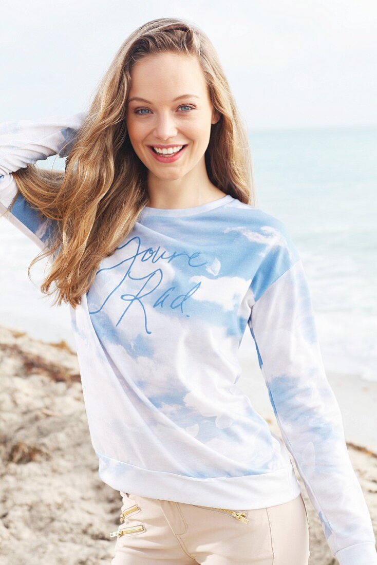A young blonde woman by the sea wearing a long-sleeved T-shirt and light trousers