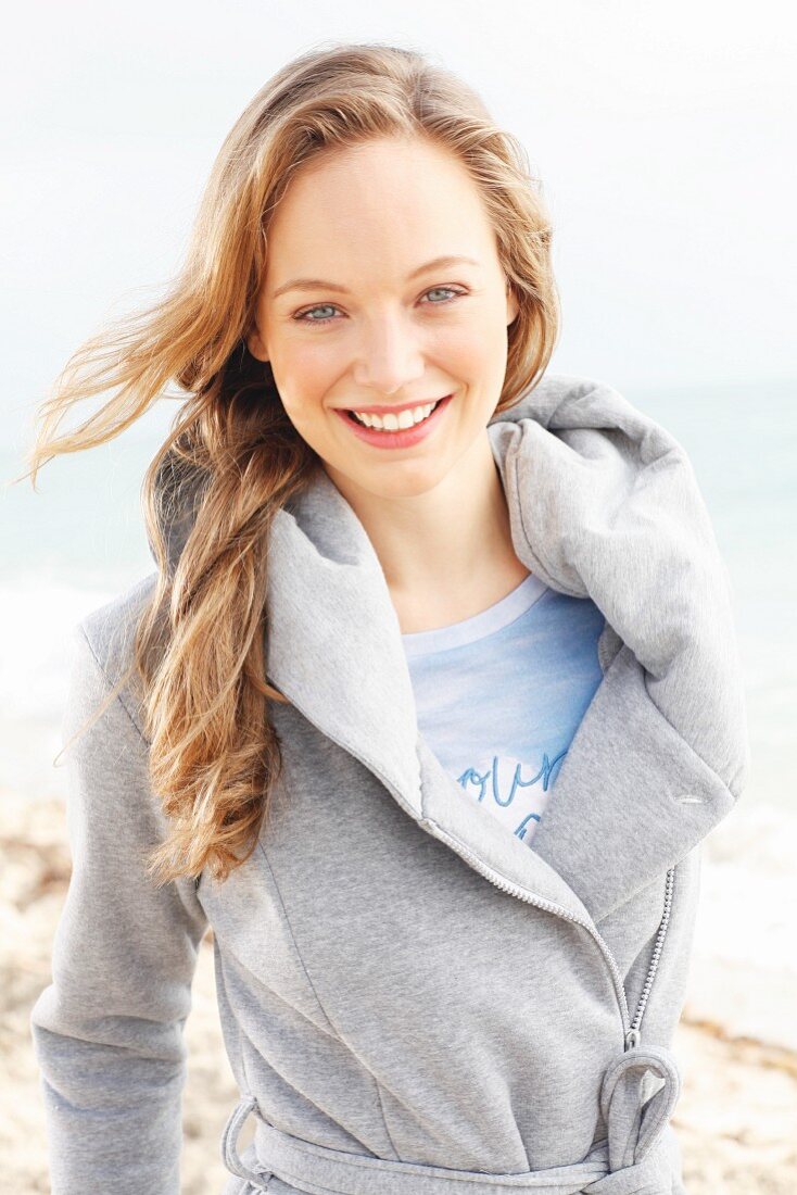 A young blonde woman by the sea wearing a grey sweatshirt coat