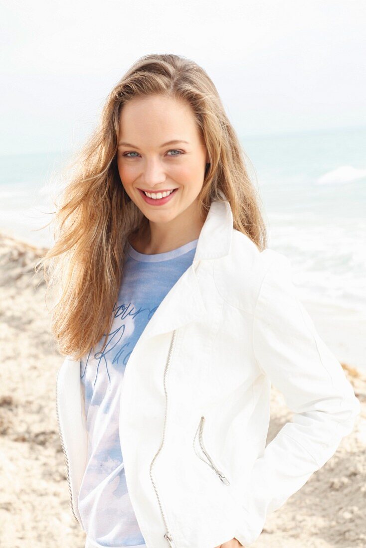 A young blonde woman on the beach wearing a shirt and a short, white jacket