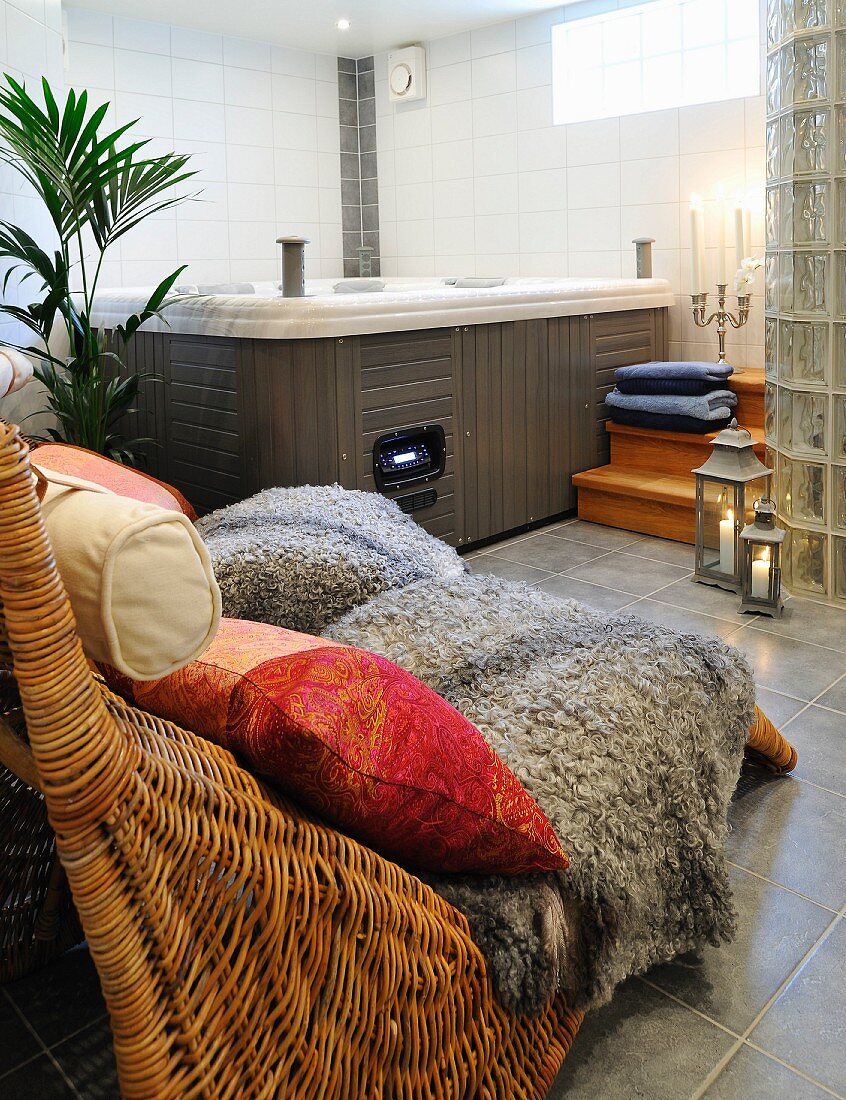 Patterned cushions and sheepskin rugs on comfortable rattan loungers in spa bathroom with whirlpool in corner