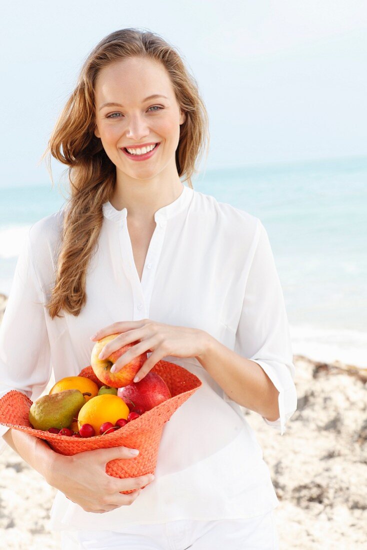 A young woman by the sea wearing a white blouse holding a hat filled with fruit