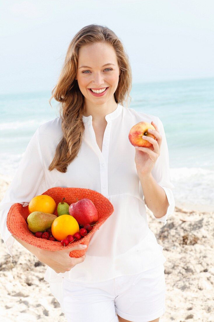 A young woman by the sea wearing a white blouse and shorts holding a hat filled with fruit