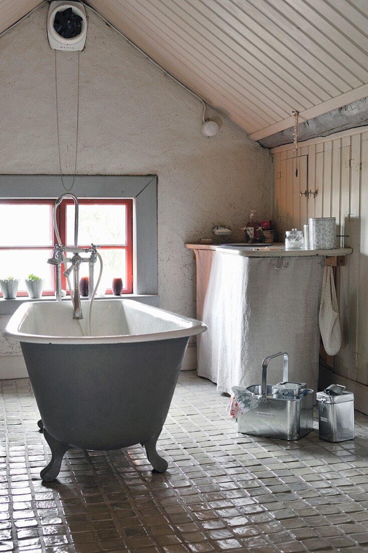 Free-standing, vintage bathtub on glazed, tiled floor in attic bathroom with window in gable wall