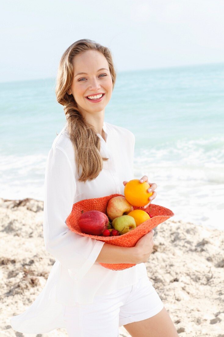 A young woman by the sea wearing a white blouse and short holding a hat filled with fruit