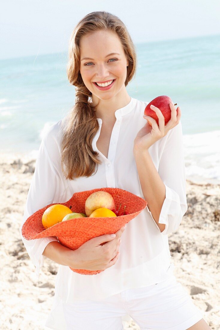 A young woman by the sea wearing a white blouse holding a hat of fruit