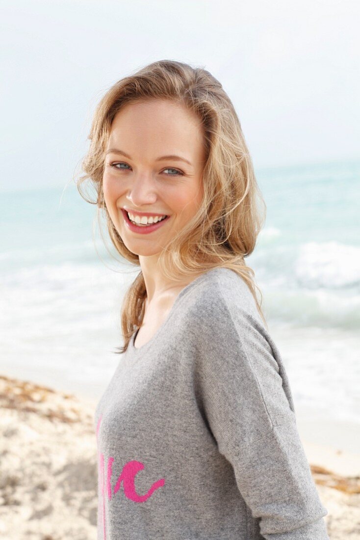 A young blonde woman on the beach wearing a grey jumper with pink writing
