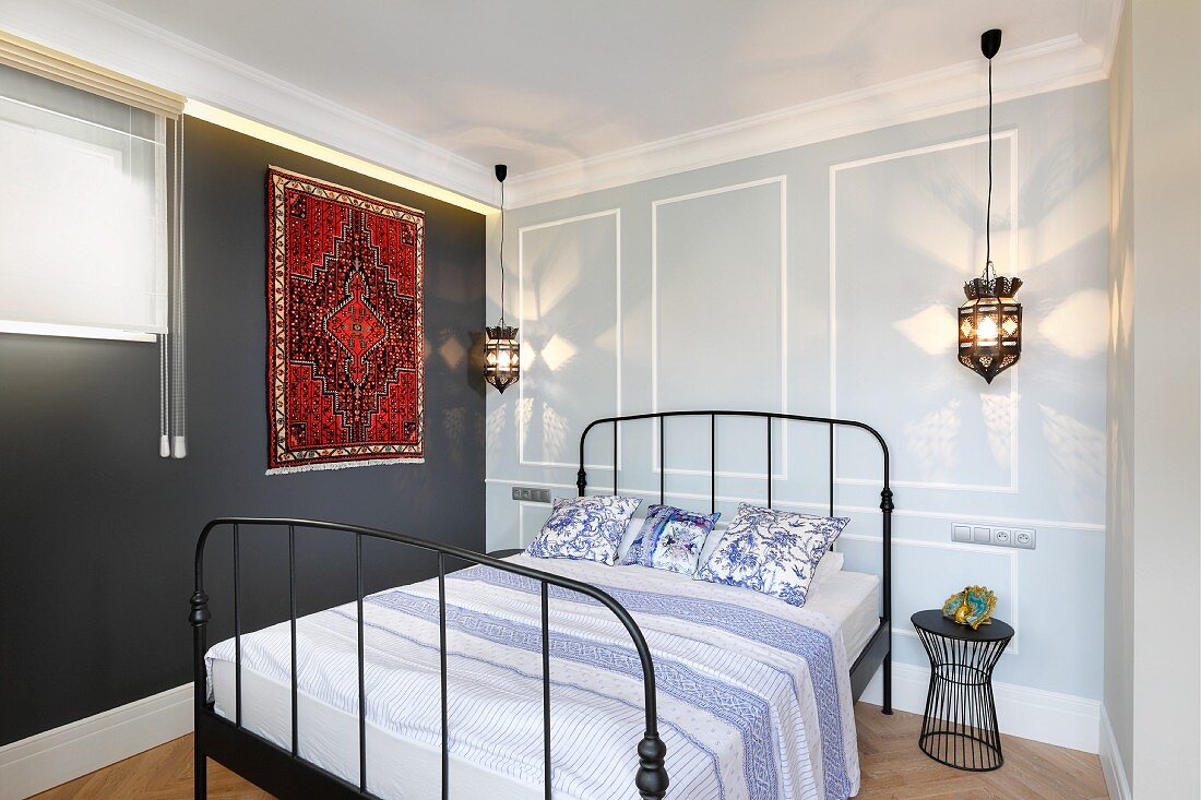 Schlafzimmer mit Doppelbett, Kopf- und Fussteil aus schwarzem Metallgitter, seitlich an dunkelgrauer Wand Teppich und Pendelleuchten in marokkanischem Stil