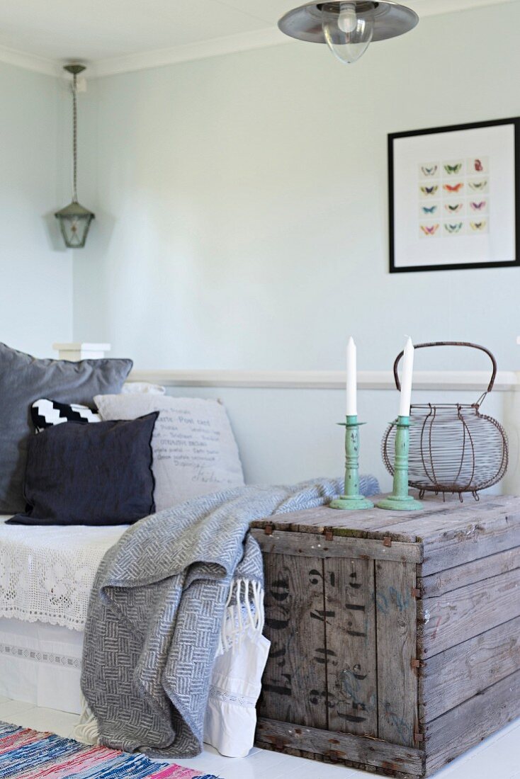 White candles in candlesticks on vintage wooden crate next to daybed in corner of simple room