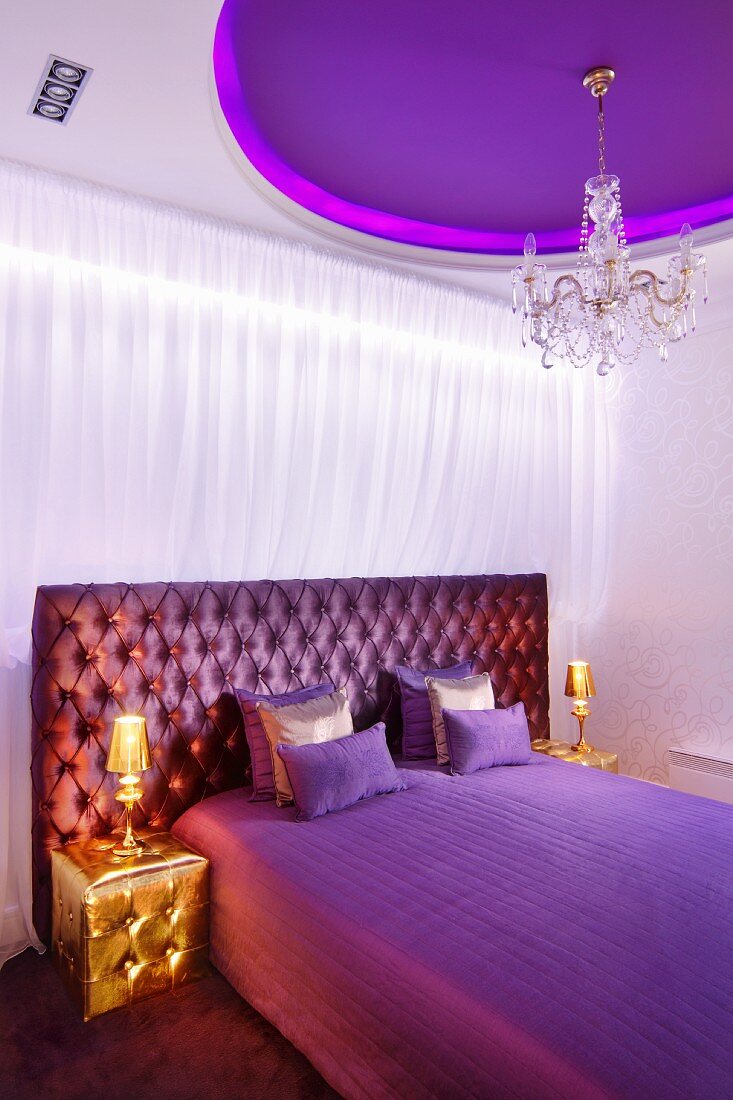 Elegantes Schlafzimmer in Violetttönen und goldfarbenen Polsterhockern mit passenden Nachttischlampen, lilafarbene Deckengestaltung und Kristallkronleuchter