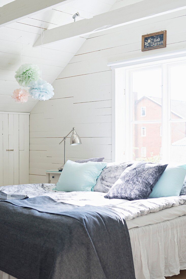 Scatter cushions and blankets on bed below window in wood-clad attic room decorated with pastel paper pompoms