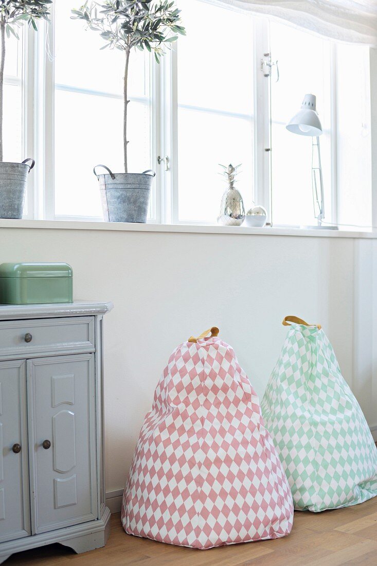 Small, pastel beanbags and retro cabinet below ribbon window with potted olive trees on windowsill