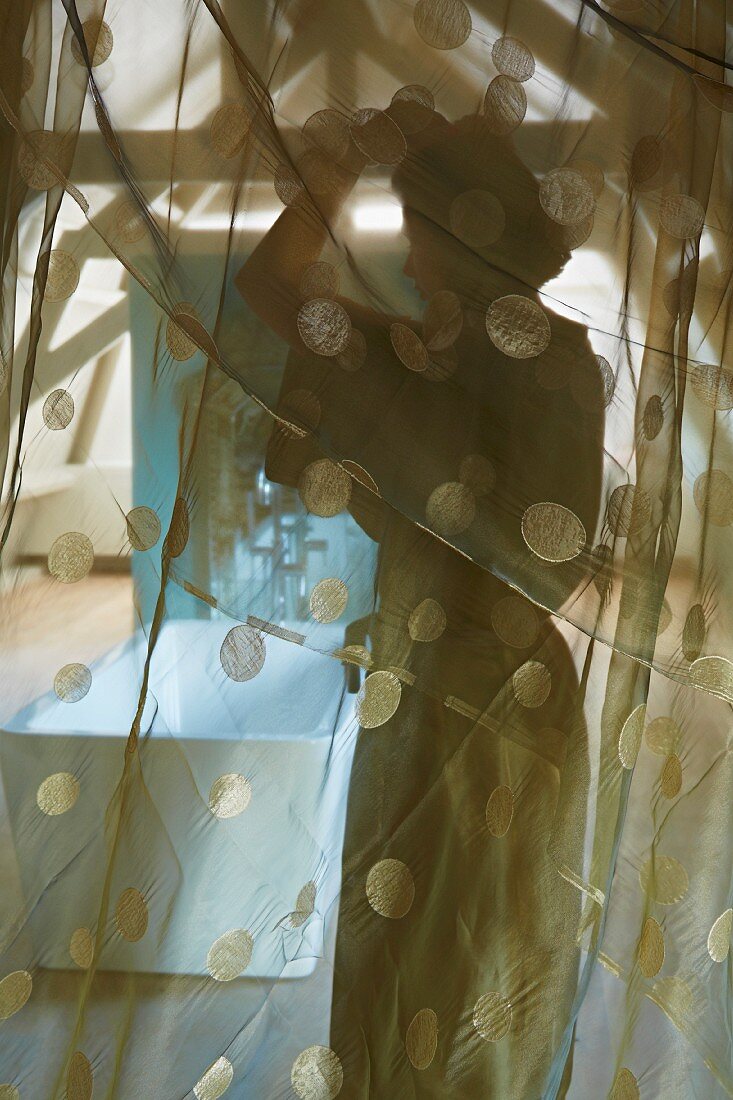 Veiled view of woman and free-standing bathtub in attic seen through translucent curtain