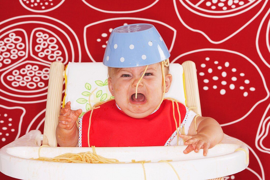 A crying baby in a high chair with a bowl of spaghetti on his head