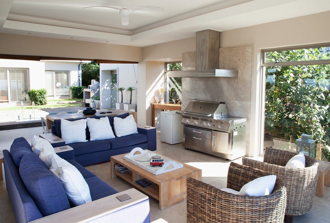 Blaue Sofas und Rattansessel in sonnigem Sitzbereich mit Grillstation, im Hintergrund Blick in den Innenhof mit Pool