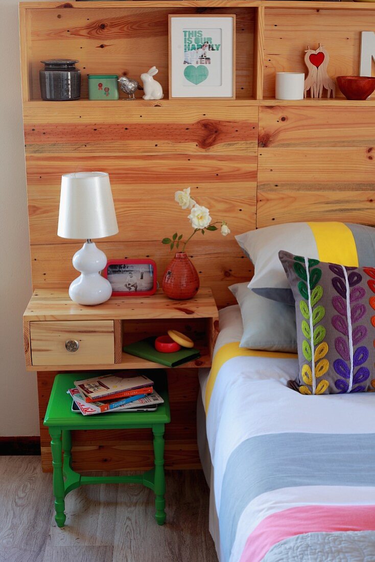 Double bed with striped bedspread, colourful scatter cushions and wooden headboard panel with youthful ornaments on integrated shelves and bedside table