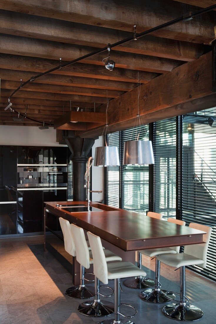 Elegant wooden counter and white swivel bar stools below pendant lamps and spotlights attached to power rail on wood-beamed ceiling; floor-to-ceiling windows with louver blinds