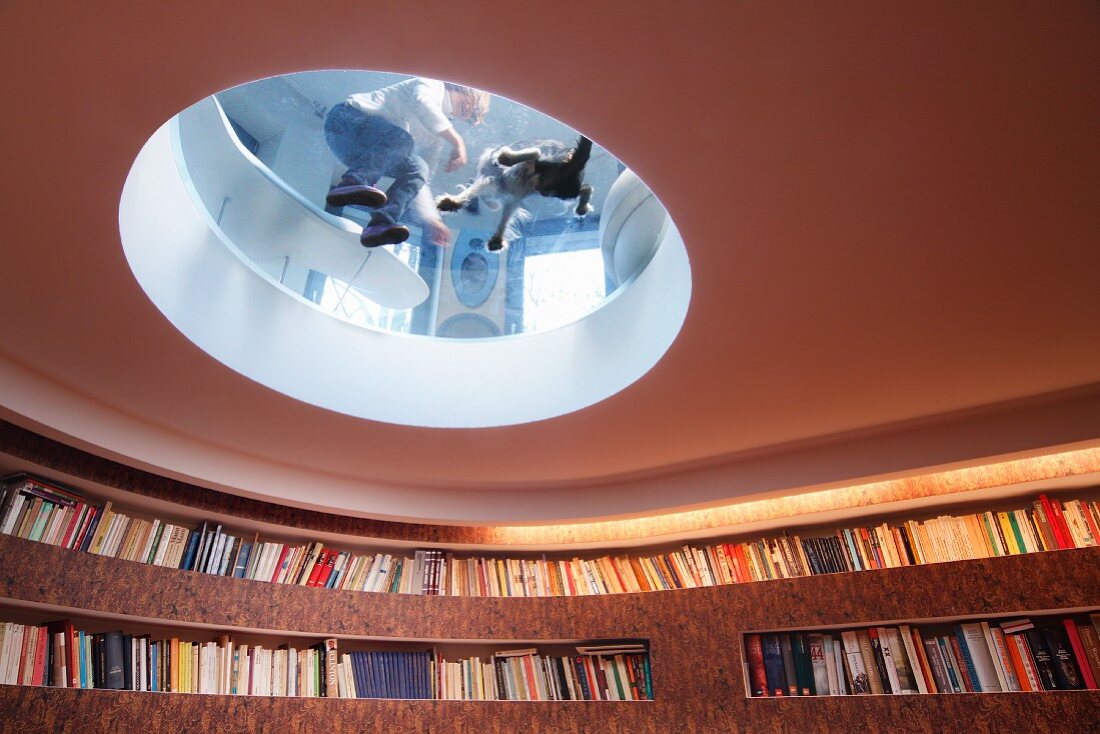 Transparente Glasplatte in kreisförmigen Deckenausschnitt mit Blick auf Kind und Hund, in Bibliothek mit gebogener Wand und Regaleinbau