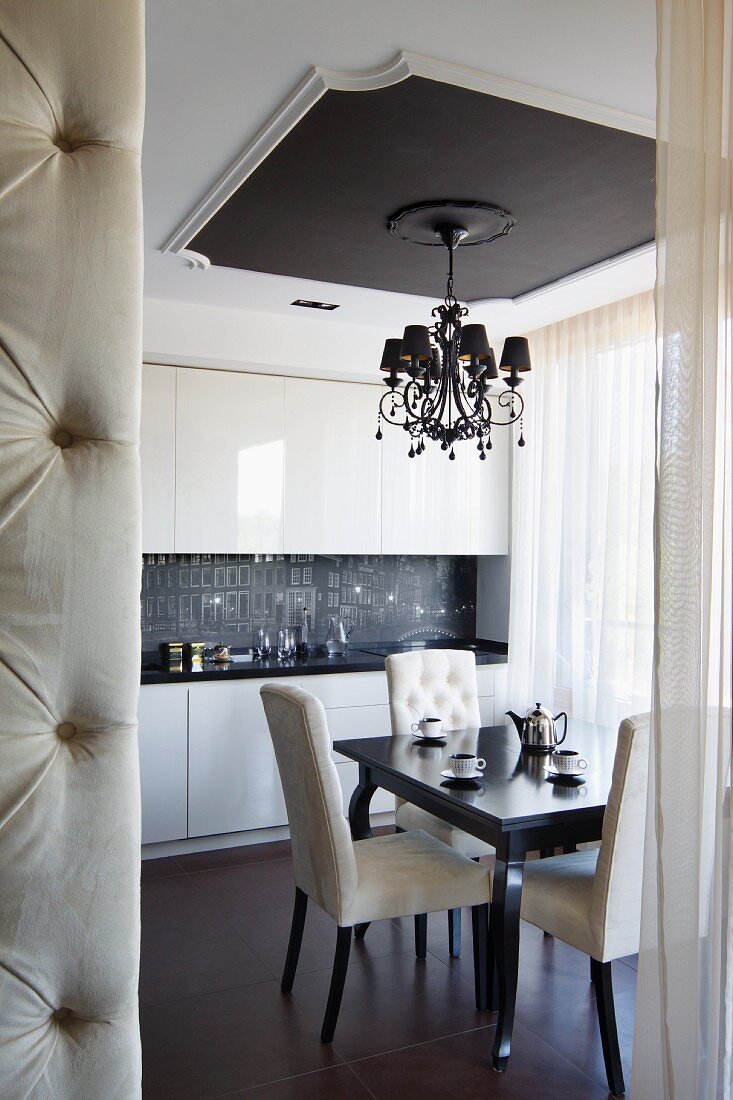 Chandelier hanging from ceiling panel in elegant kitchen-dining room; edge of button-tufted partition in foreground