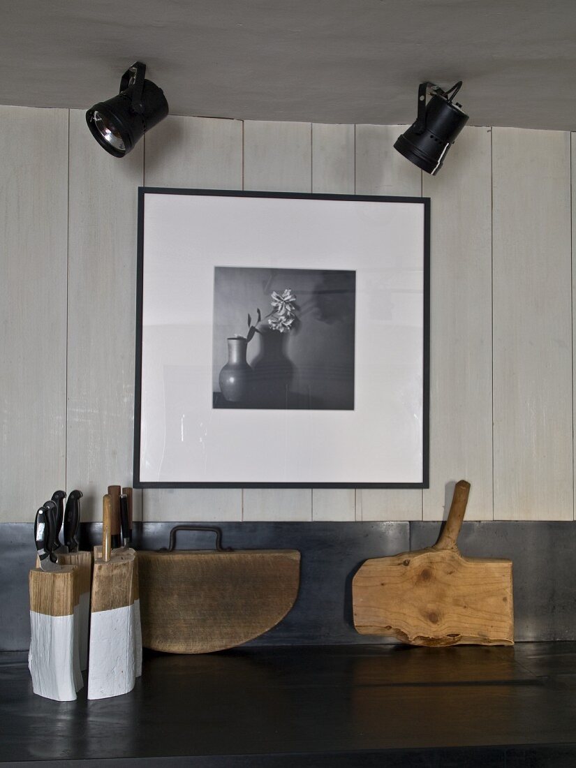 Knife block and rustic chopping boards on kitchen worksurface below ceiling spotlights and black and white photo on white wooden wall