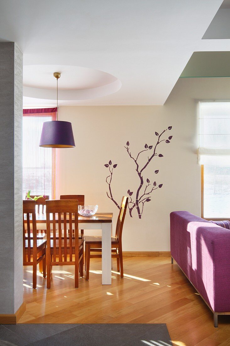 Elegant dining area with wooden chairs below purple pendant lamp, stencilled branch motif on wall and couch to one side