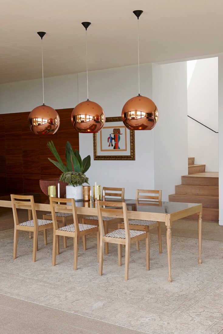 Long designer dining table and chairs made from pale wood under copper-coloured lampshades in modern dining area