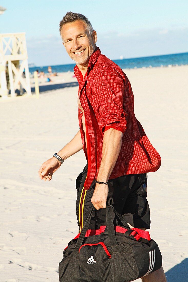 A middle-aged man on a beach wearing a red shirt and bathing shorts holding a sports bag