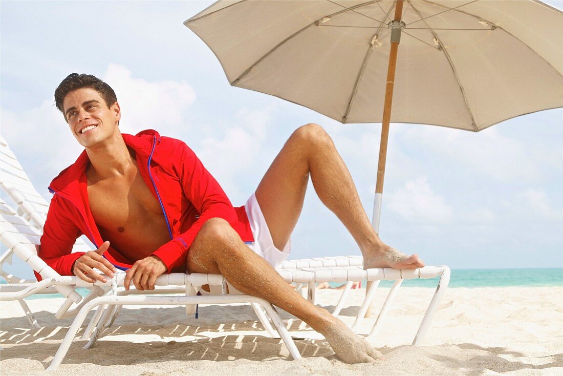 Junger Mann mit roter Kapuzenjacke und Shorts auf Sonnenliege am Strand