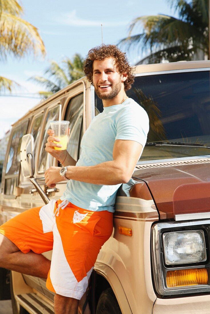 A young man with curly hair and a beard wearing summer clothes leaning against a car