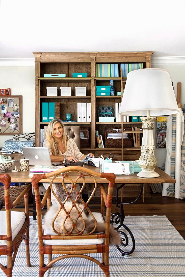 Woman seated at desk, large table lamp with white lampshade, bamboo chairs and traditional wooden shelves of office supplies