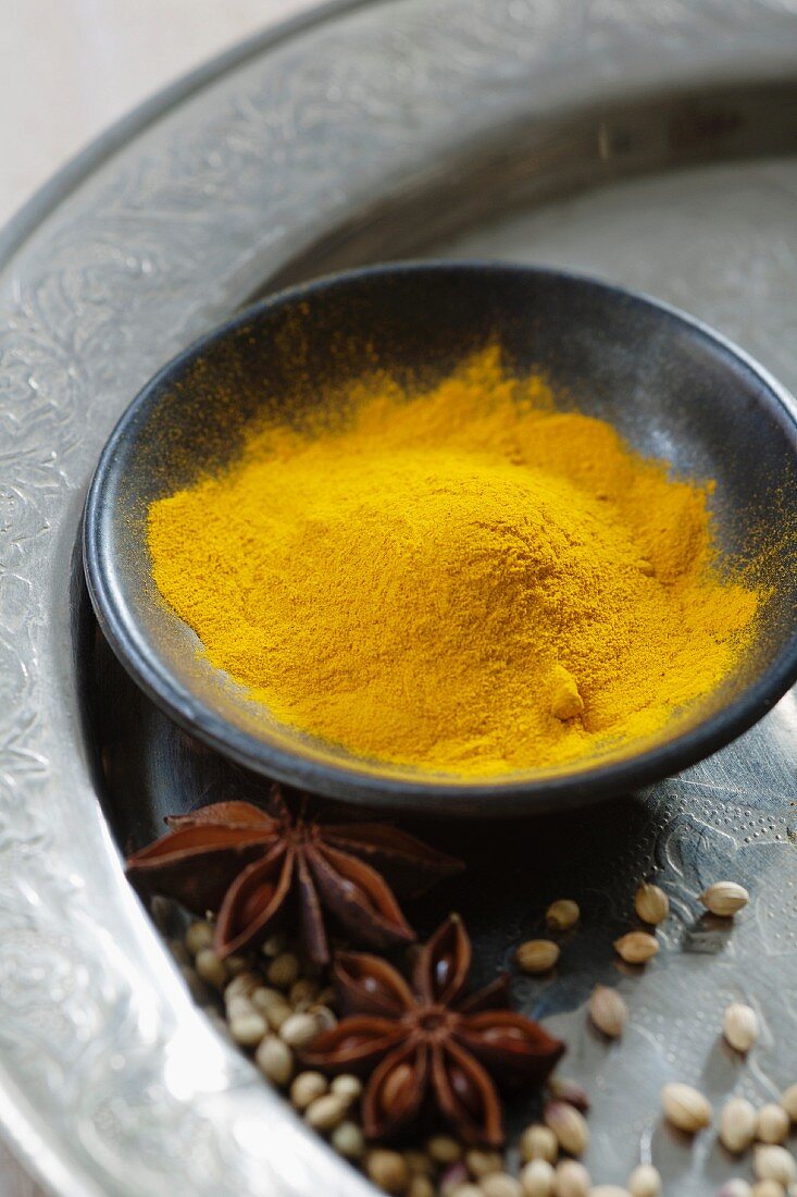 Turmeric powder in a black bowl with star anise and coriander on a silver plate