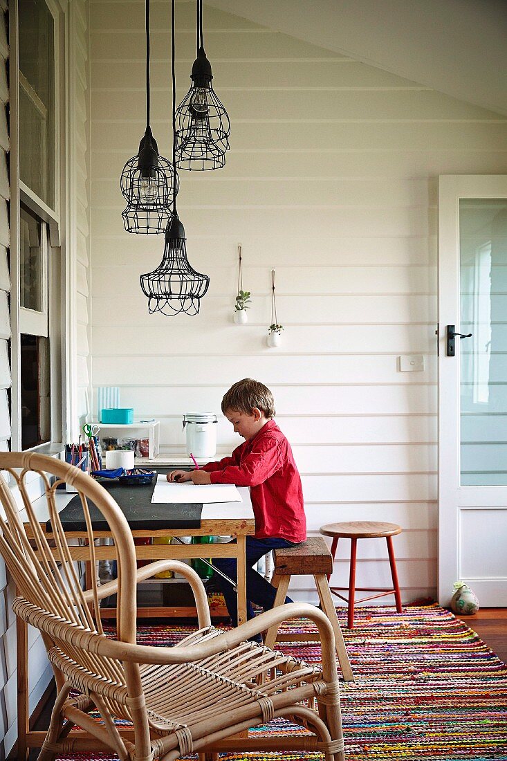 Kind am Schreibtisch und verschiedene Pendelleuchten mit Drahtlampenschirmen, im Vordergrund Rattanarmlehnsessel
