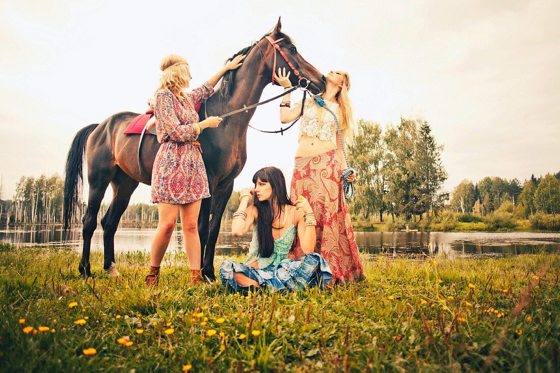 Three young women wearing hippie-style clothing with a horse by a lake