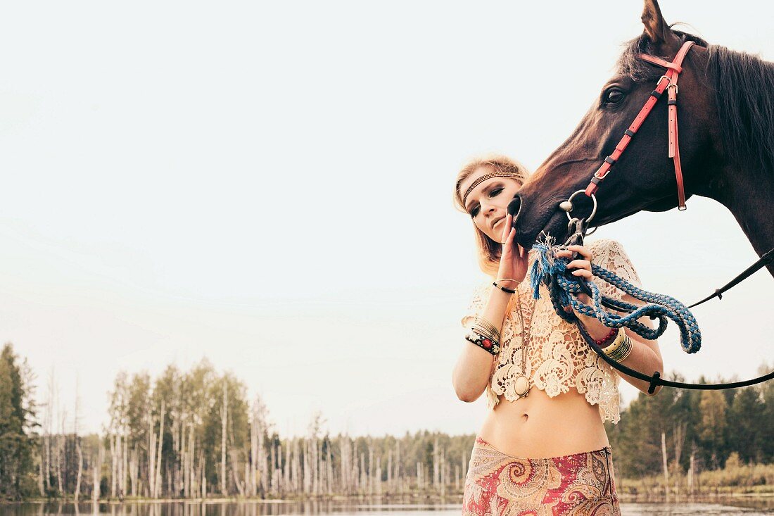 A young blonde woman wearing hippie-style clothing stroking a horse by a lake