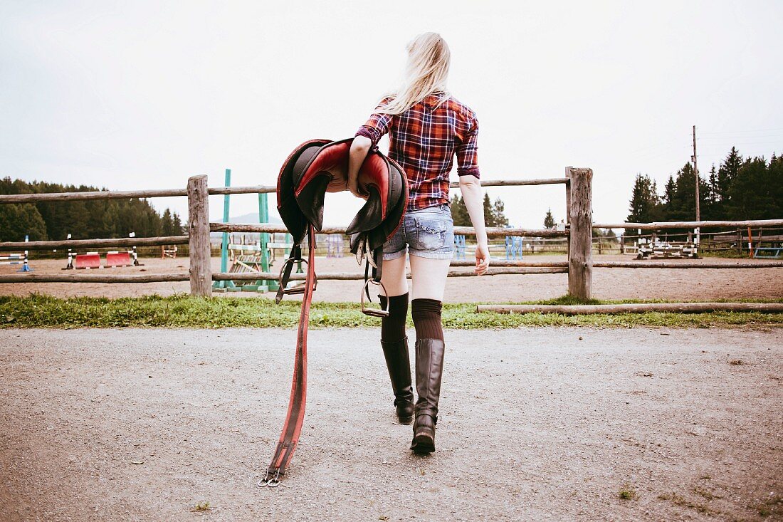 A long-haired blonde woman wearing a checked shirt and short shorts carrying a saddle