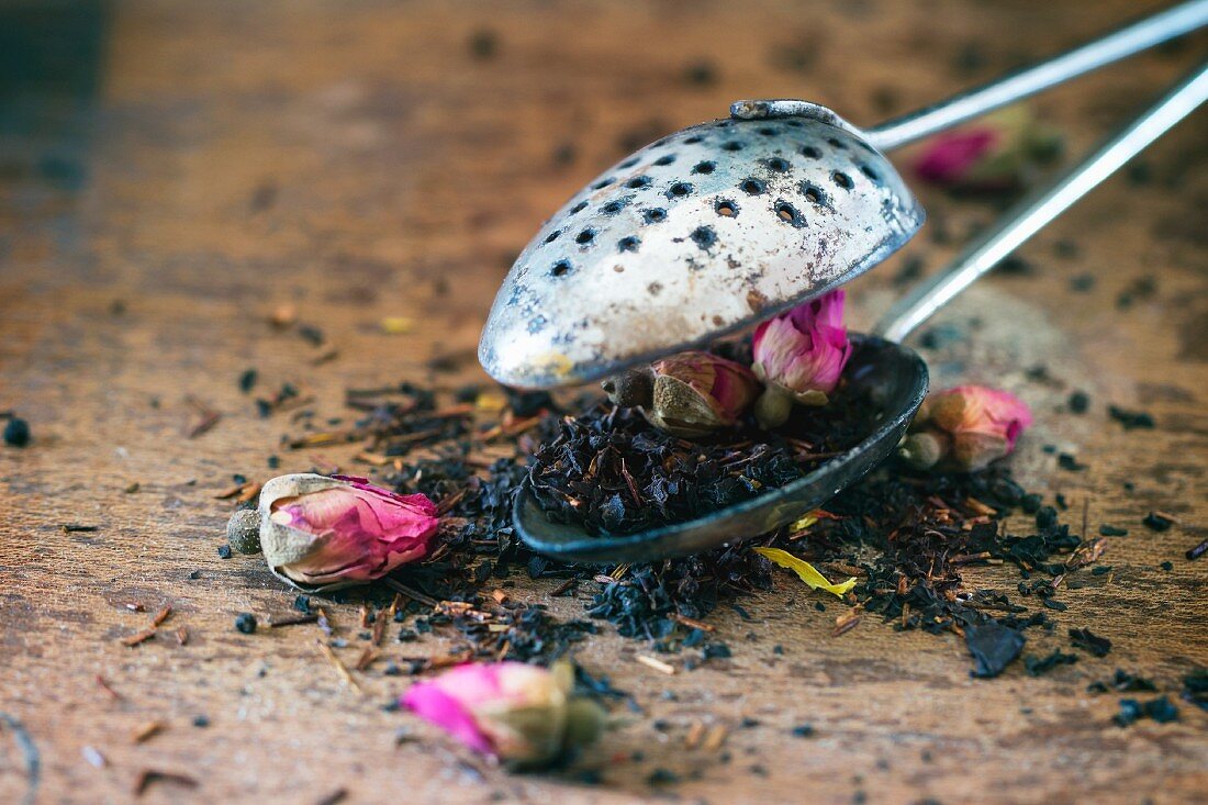 Getrocknete Teeblätter mit Rosenknospen im Vintage-Teesieb auf Holzuntergrund