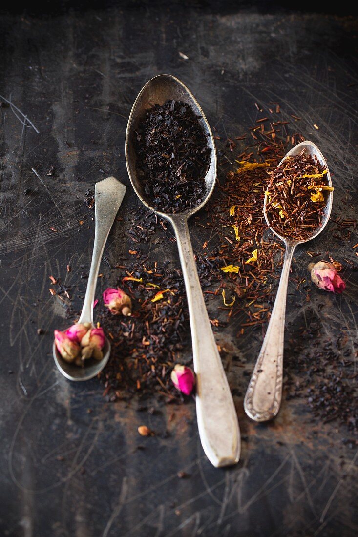 Getrockneter schwarzer Tee und Rooibos mit Rosenknospen im Vintage-Löffel auf Metalloberfläche