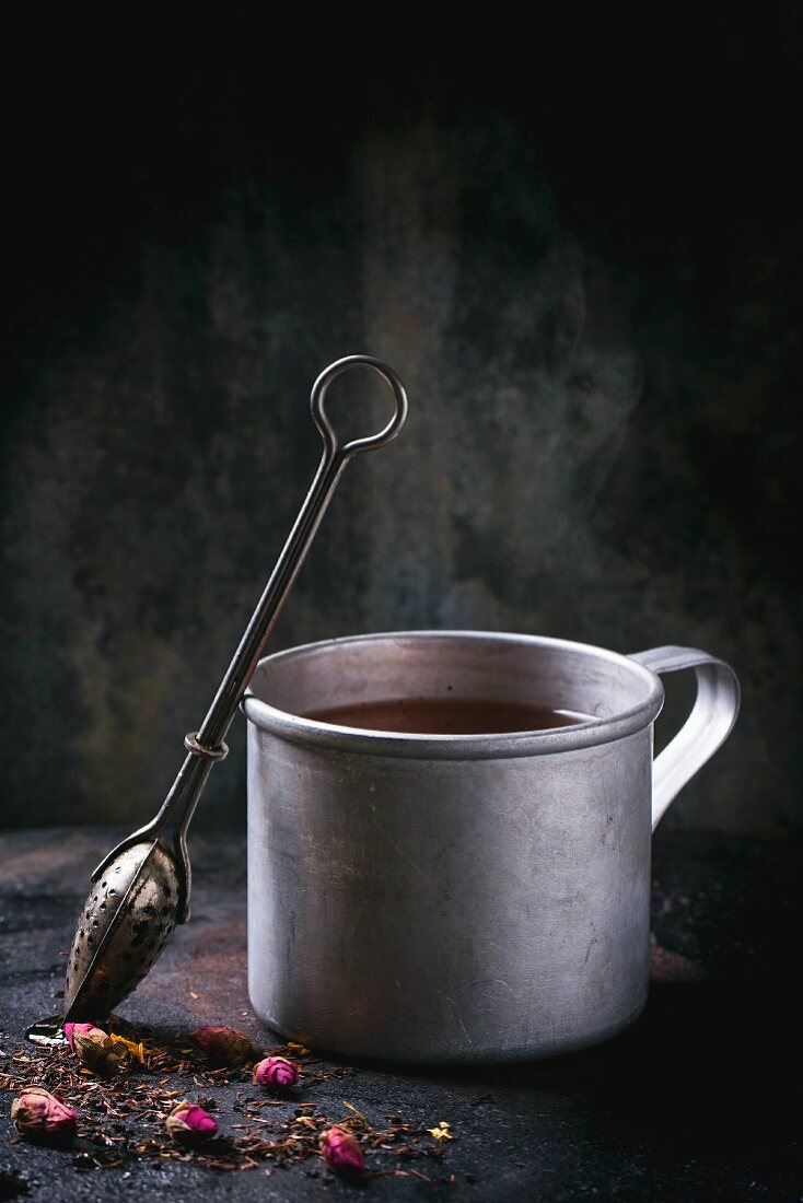 Tee in Blechtasse mit Teesieb und getrockneten Rosenknospen auf Metalloberfläche