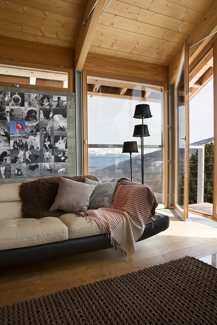 Ledersofa vor Wand mit Fotosammlung, seitlich Stehleuchtengruppe vor Panoramafenster, in holzvergekleidetem Chalet