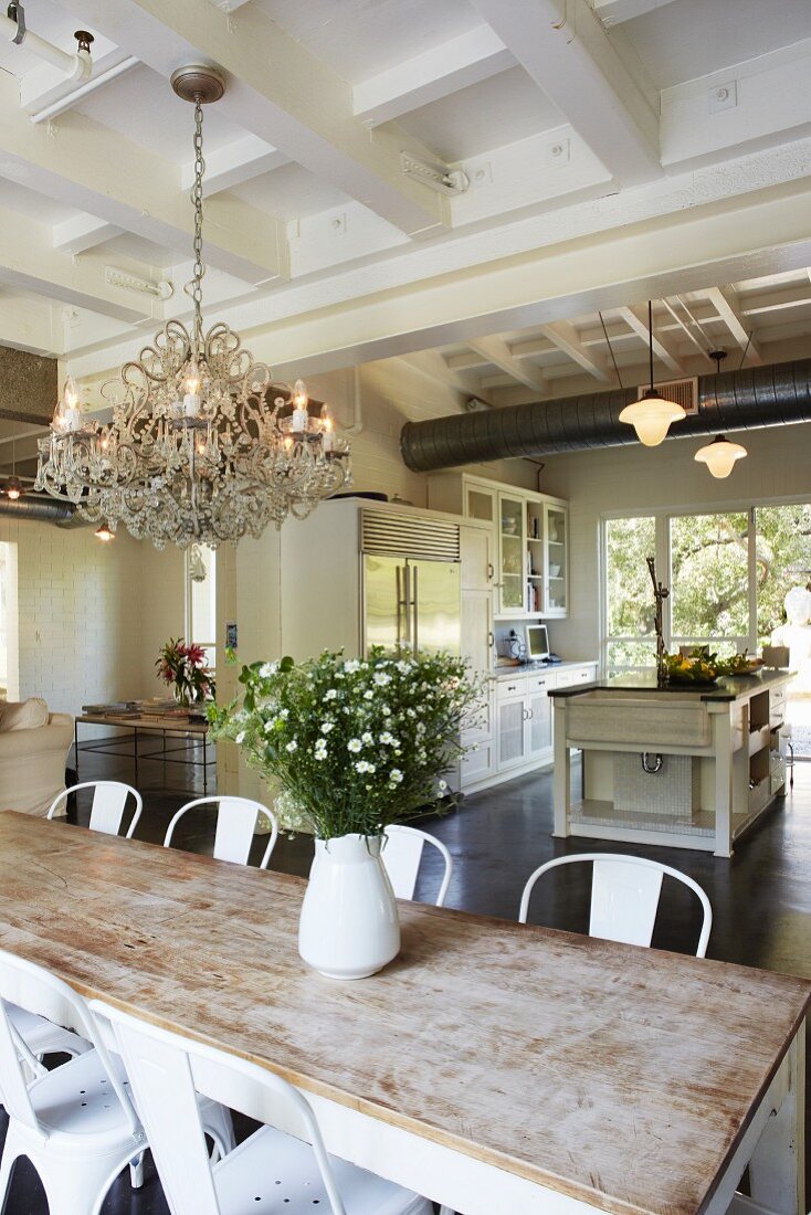 Dining area with classic chairs around rustic table, white jug of garden flowers and open-plan kitchen in background