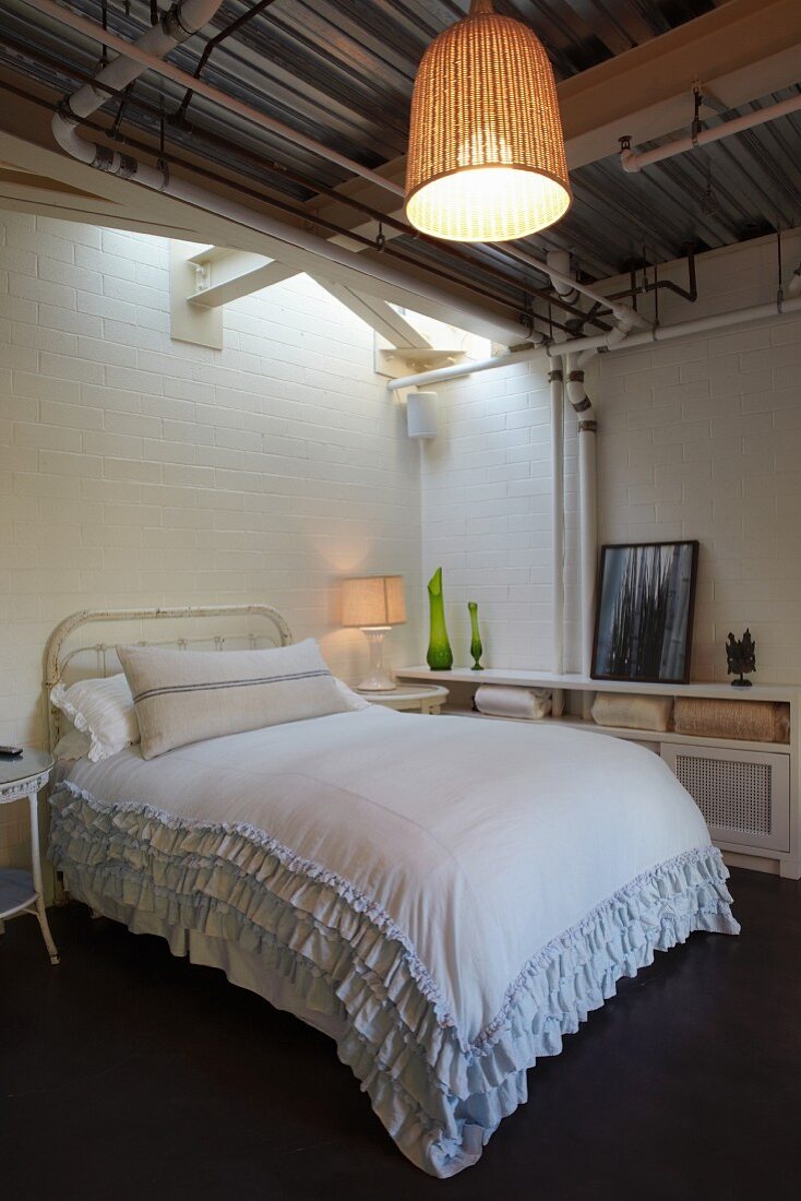 Double bed with ruffled bedspread in simple room with wicker pendant lampshade hung from corrugated metal ceiling