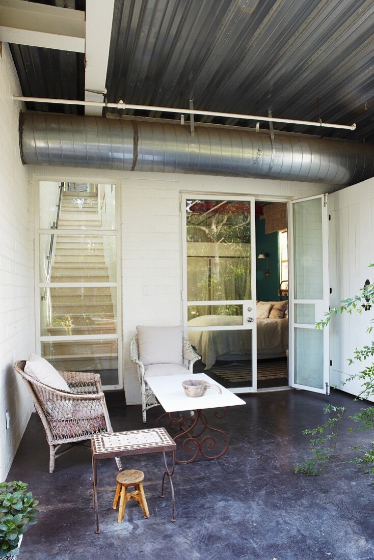 Terrace furnished with wicker chair, stool and small table outside contemporary house with ventilation duct running below corrugated metal ceiling