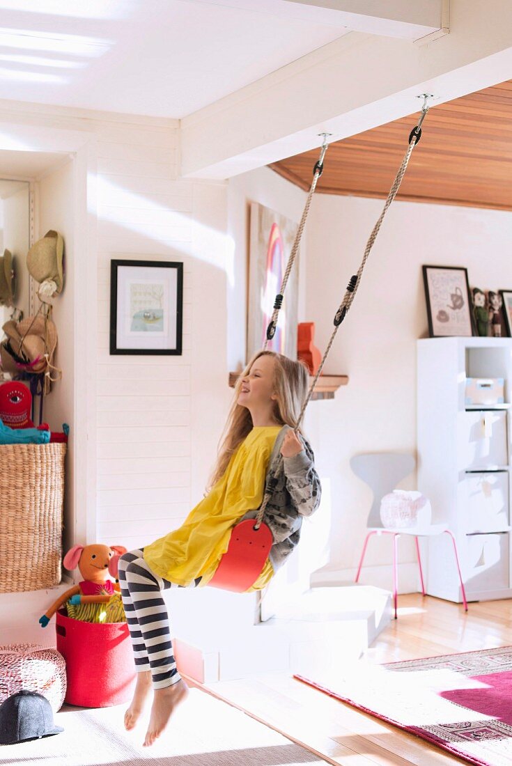 Girl on swing in open-plan interior, toys on floor and retro chair and shelving next to open doorway